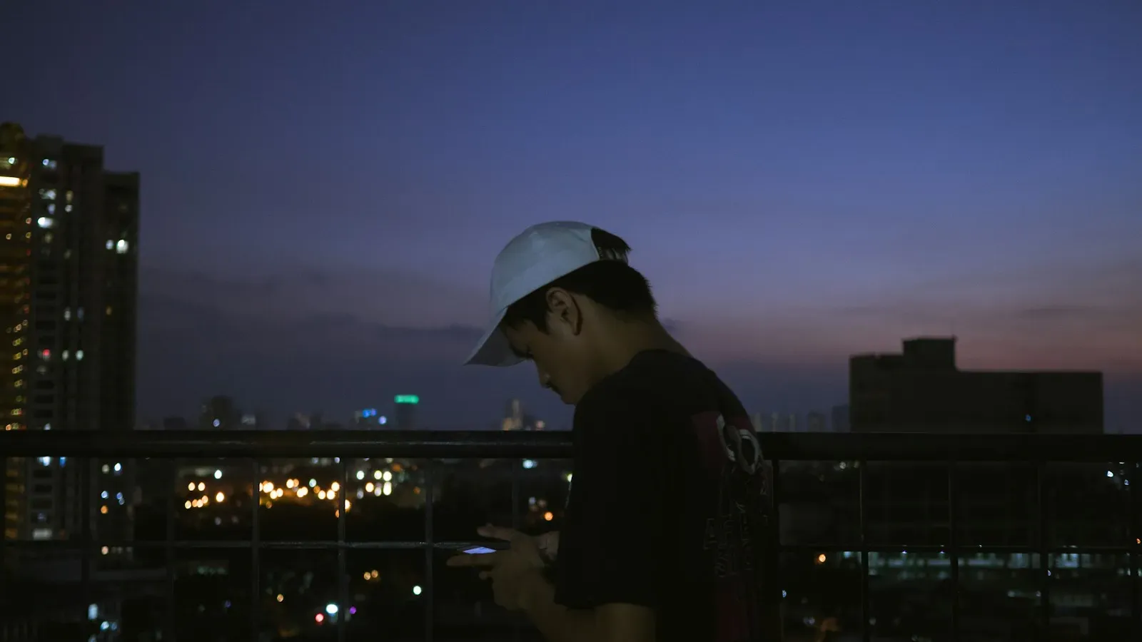 Mann steht nachts auf einem Balkon und schaut auf sein Smartphone – sicher im Gay Chat
